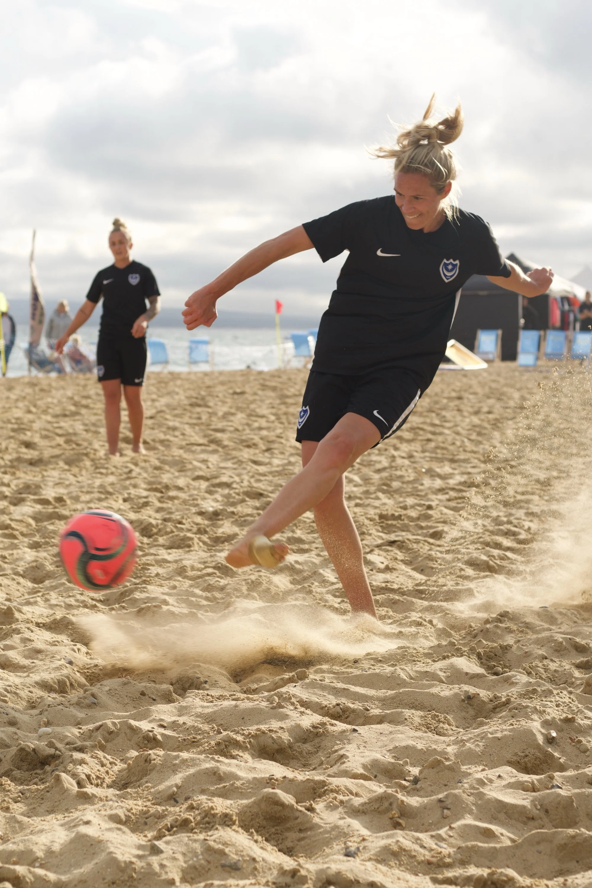Women playing beach soccer on sandy beach, one kicking pink and black soccer ball, others watching, cloudy sky.