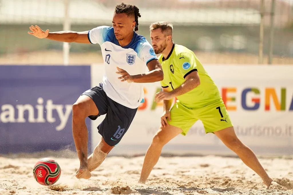 Two men playing soccer on a sandy field. One man, wearing a white and blue jersey, is kicking a red and black soccer ball. The other man, in a yellow jersey, is trying to block or defend. Both players are focused on the ball, with a background of advertising boards and a slightly blurred outdoor setting.