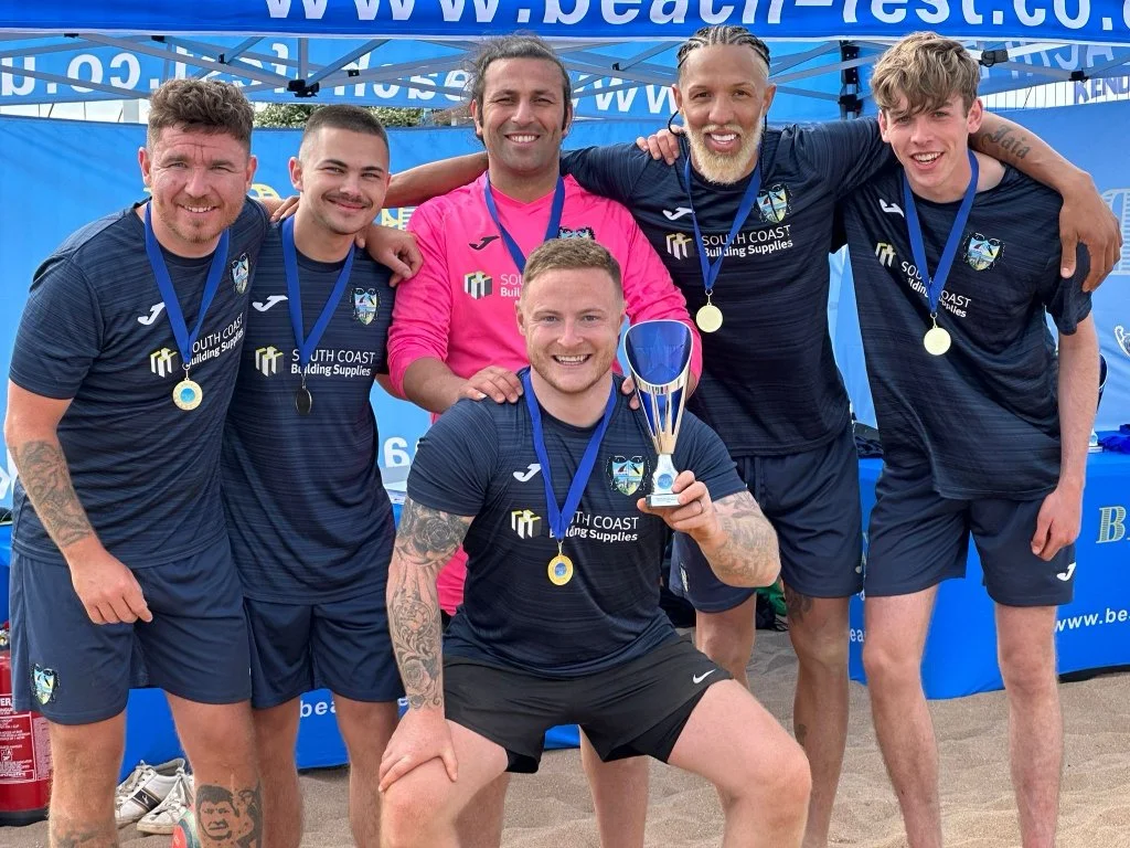 Six men in matching navy sports uniforms celebrating with medals and a trophy in front of a blue tent at a beach event.