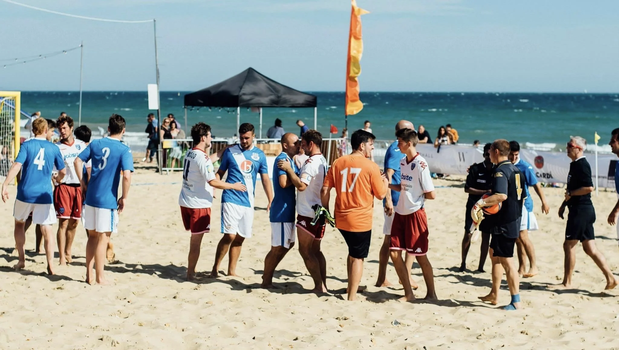 Group of soccer players on a sandy beach after a game, some wearing white and maroon uniforms, others in blue and white, shaking hands and chatting. The ocean and a blue sky are in the background, with a tent and some onlookers nearby.