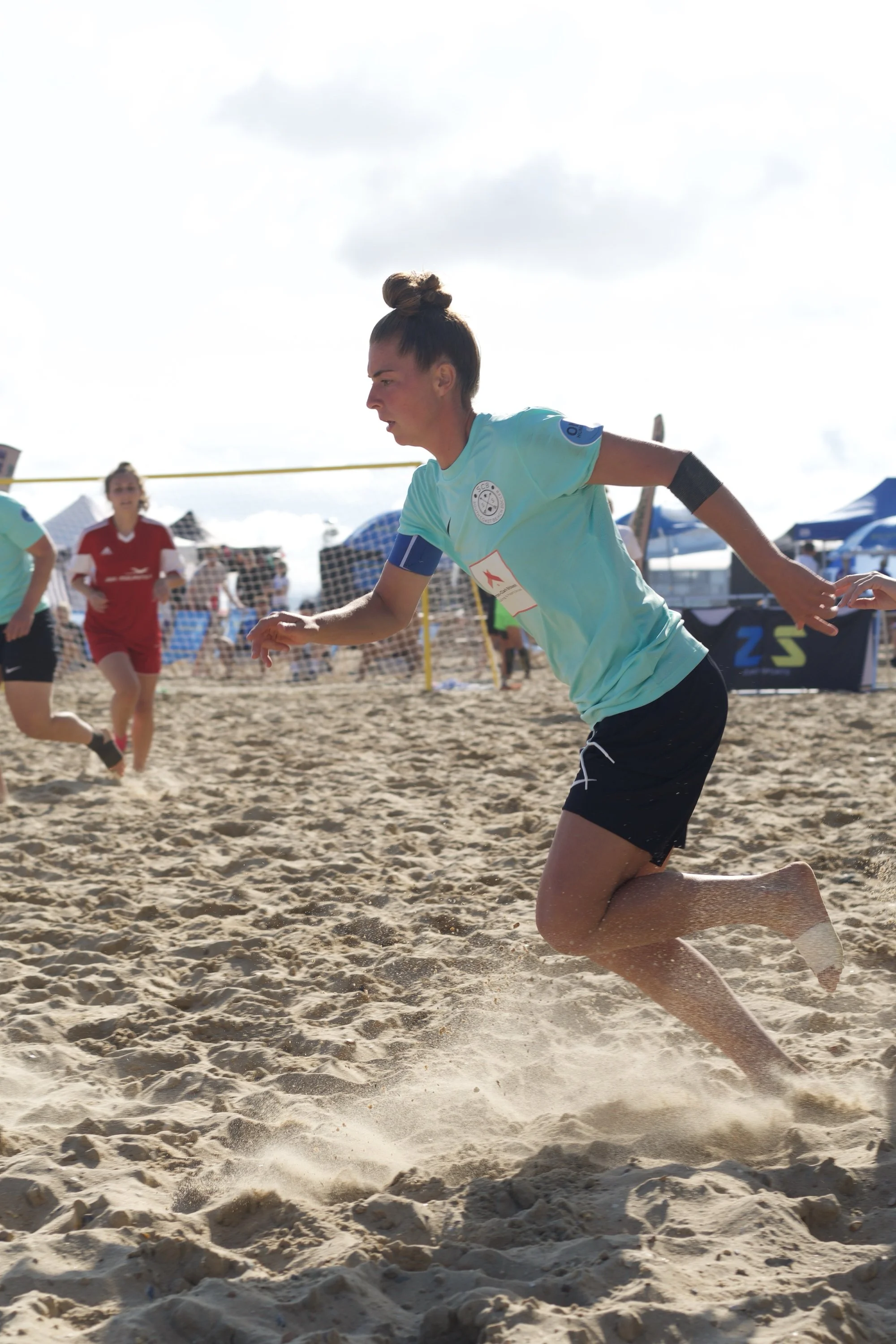 A woman playing beach soccer on sand, running with other players and sand flying around her, under a partly cloudy sky.