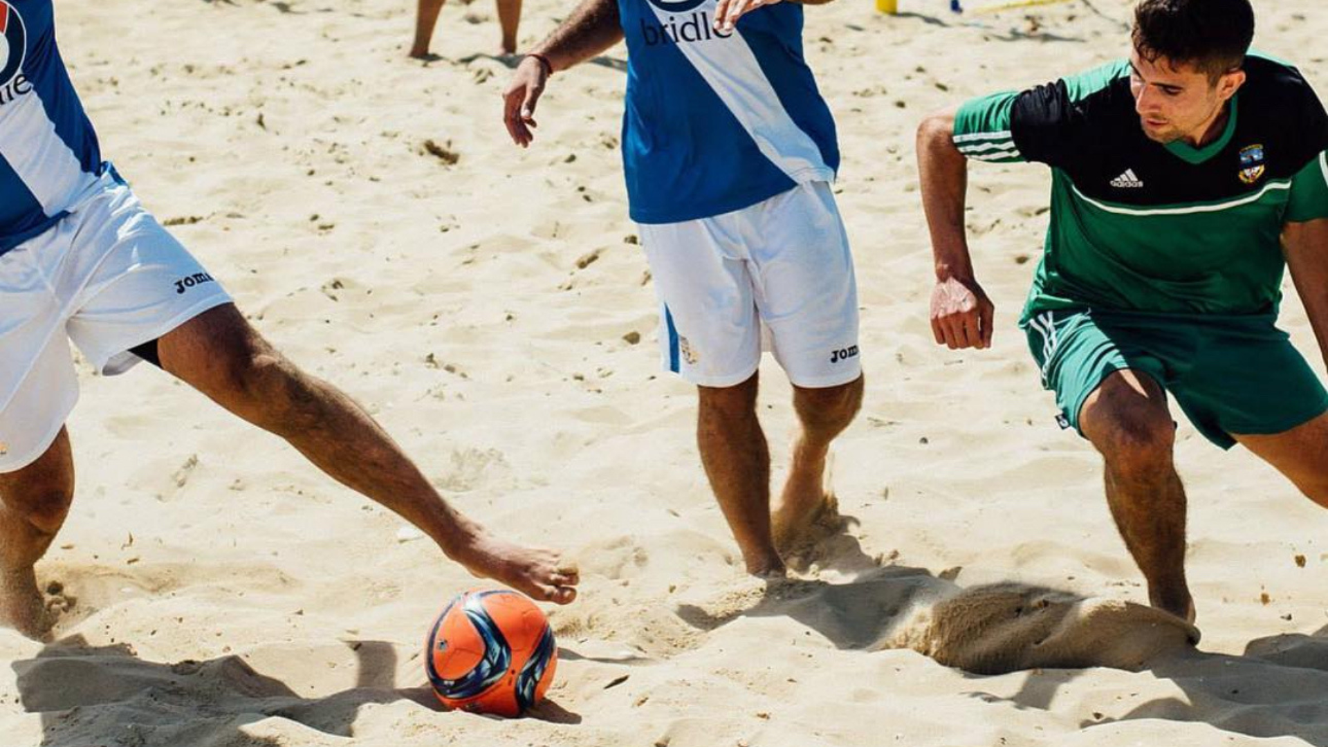 People playing beach soccer on sand, with a ball near their feet.