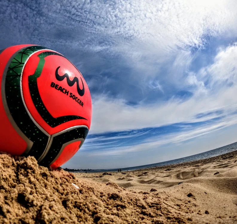 A beach volleyball resting on sandy beach with ocean and cloudy sky in the background.