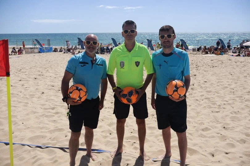Three men standing on a sandy beach holding soccer balls, with beachgoers, umbrellas, and the ocean in the background.