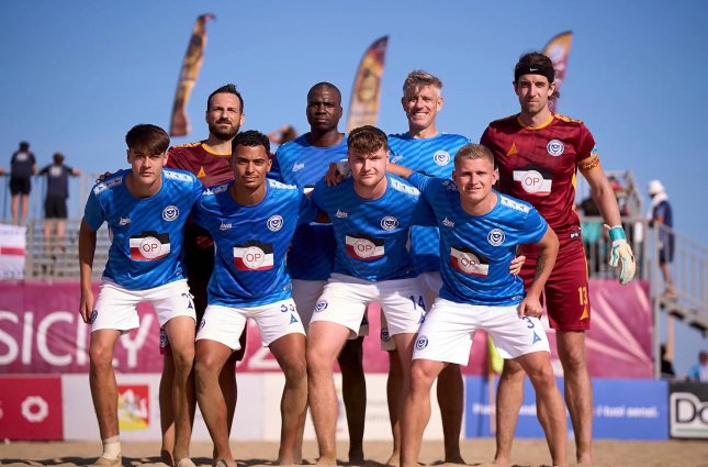 Group of seven young men in sports uniforms on a beach volleyball court, with some in blue jerseys and others in maroon, posing for a team photo under a clear blue sky.