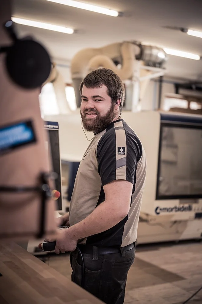 Ein lachender Mann mit dunklem Haar und Bart arbeitet an einer Maschine in einer Werkstatt.