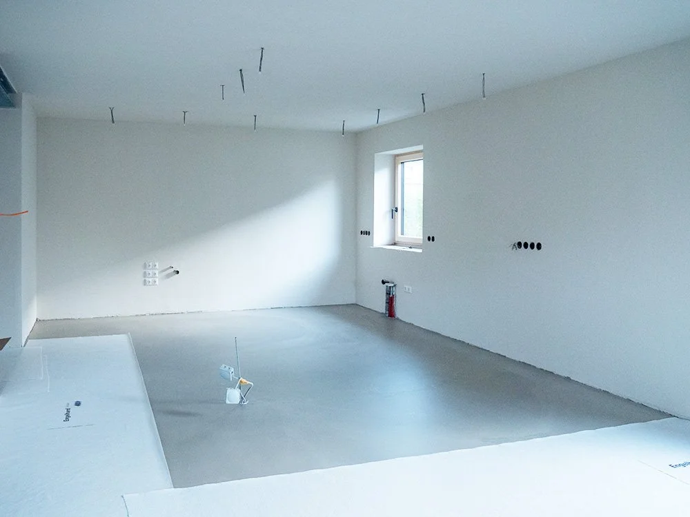 Leeres, unfertiges Zimmer mit weißen Wänden, einer kleinen Fensterfront, einigen Steckdosen, Kabeln an der Decke und auf dem Boden, und einem Feuerwehrlöscher in der Ecke.