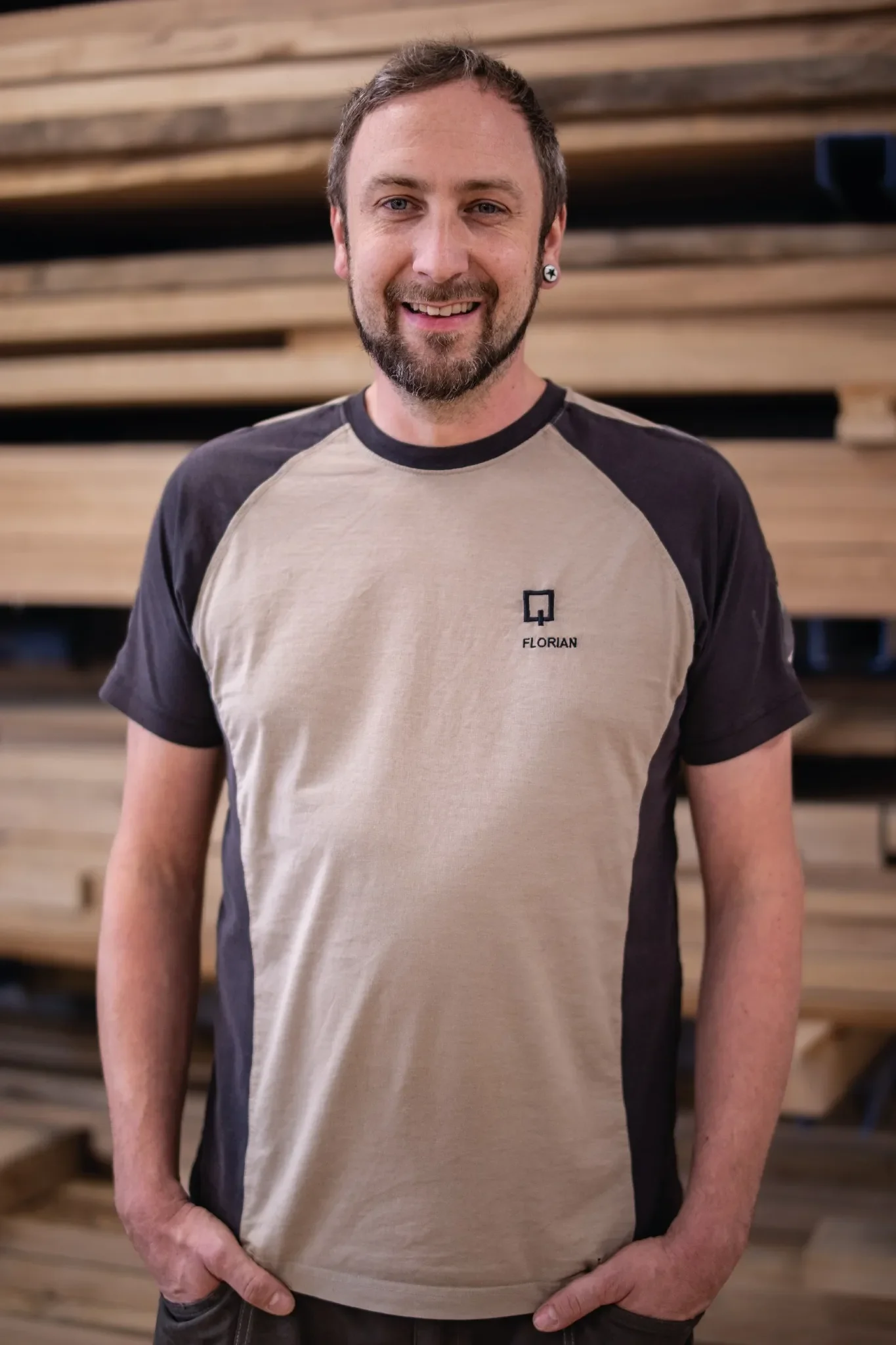 Halb-Körper-Portrait von Florian im Holzquadrat T-Shirt vor aufeinander gestapelten Brettern