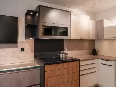 Küchenzeile mit schwarzen, weißen und Holz-Schrankfronten, Backofen, Kochfeld, Wasserkocher und Fernseher an der Wand.