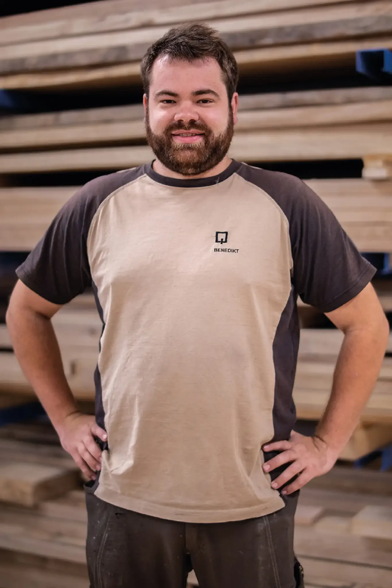 Halb-Körper-Portrait von Benedikt im Holzquadrat T-Shirt vor aufeinander gestapelten Brettern