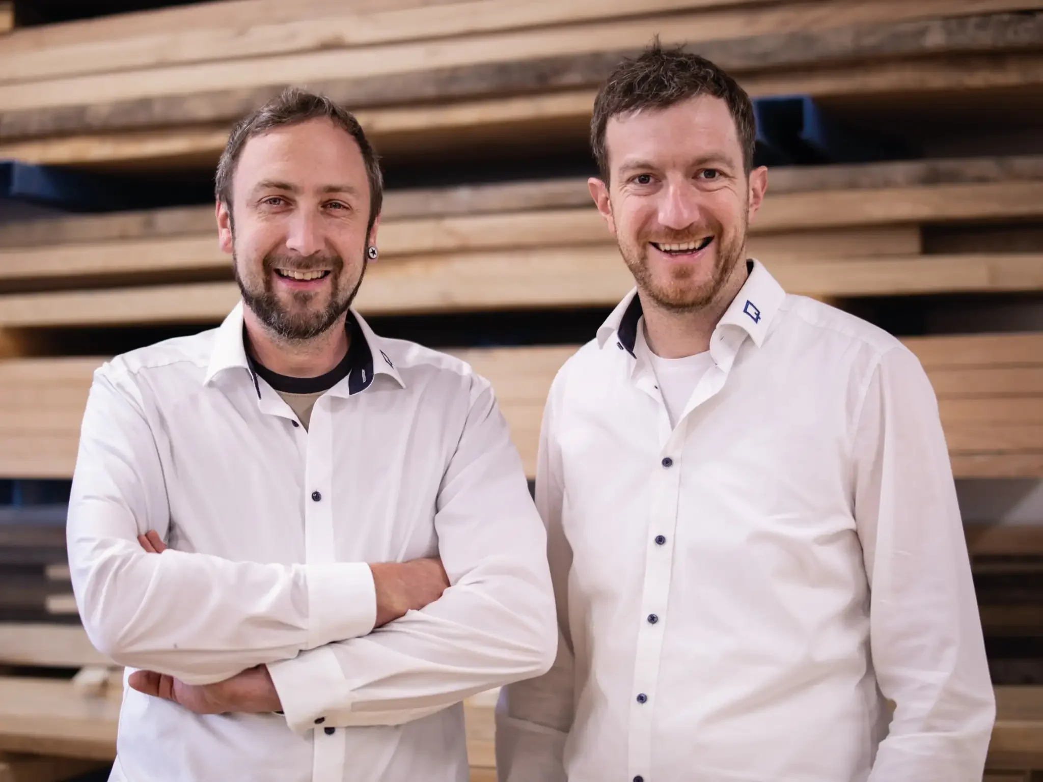 Zwei lachende Männer (Florian und Dominik) in weißen Hemden stehen vor einem Holzbstapel.