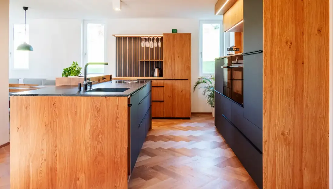 Moderne Küche mit Holz- und dunkelgrünen Schrankfronten, Kücheninsel, zwei Fenster, Pflanzen, Wein-Gläser in einem Schrank mit Glas.