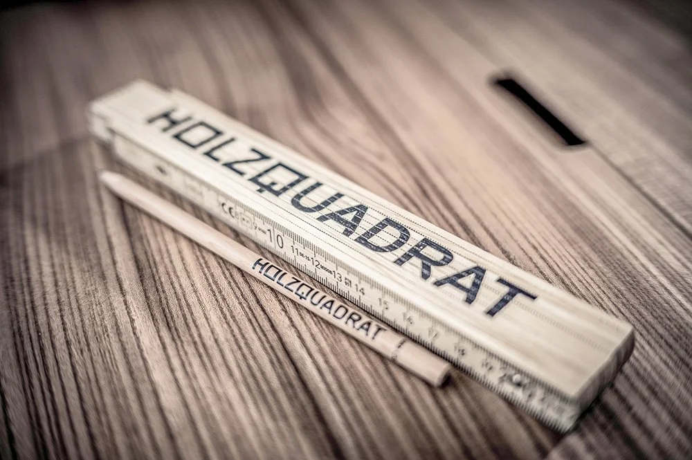 Ein Zollstock / Meterstab mit der Aufschrift "HOIZQUADRAT" liegt auf einem Holztisch.