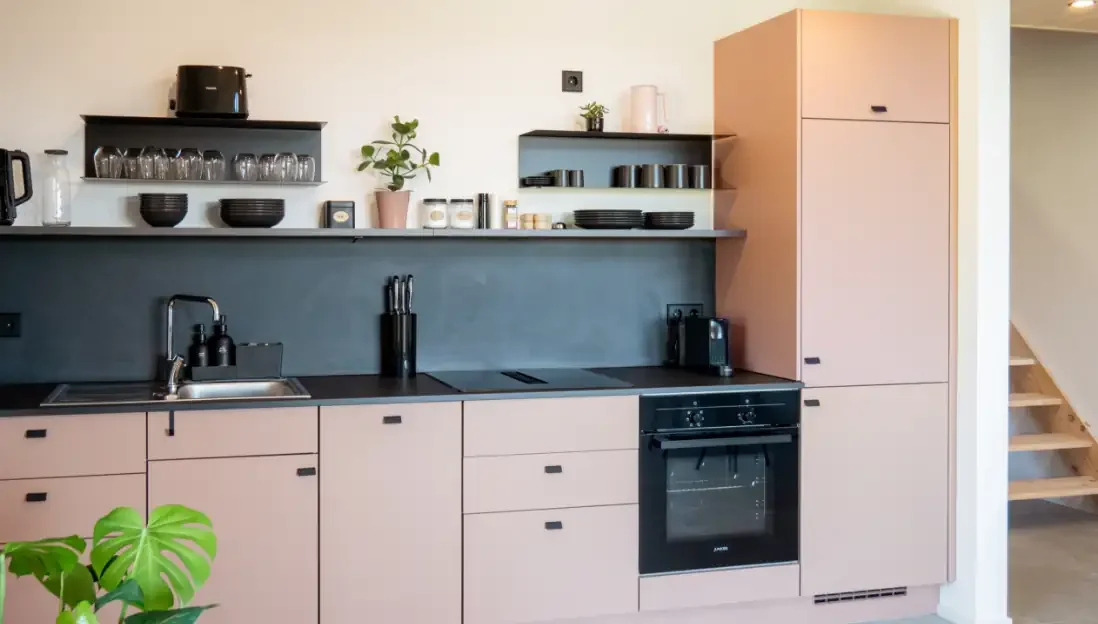 Moderne Küchenzeile mit schwarzer Arbeitsplatte, offenem Regal mit Geschirr, Küchenutensilien. Fronten sind in einem sanften Rosa gehalten. Es gibt eine Spüle, einen Herd, eine Kaffeemaschine und einem Messerblock.