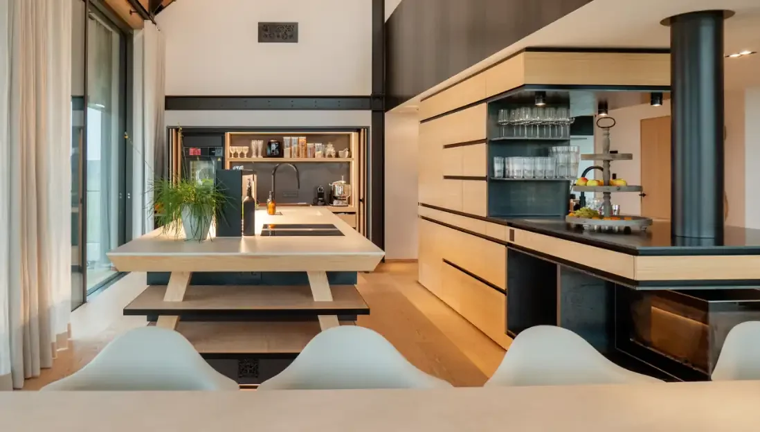 Moderne Küchenansicht mit Esstisch, Küchenregalen, Küchengeräten und Fenstern mit Vorhängen.