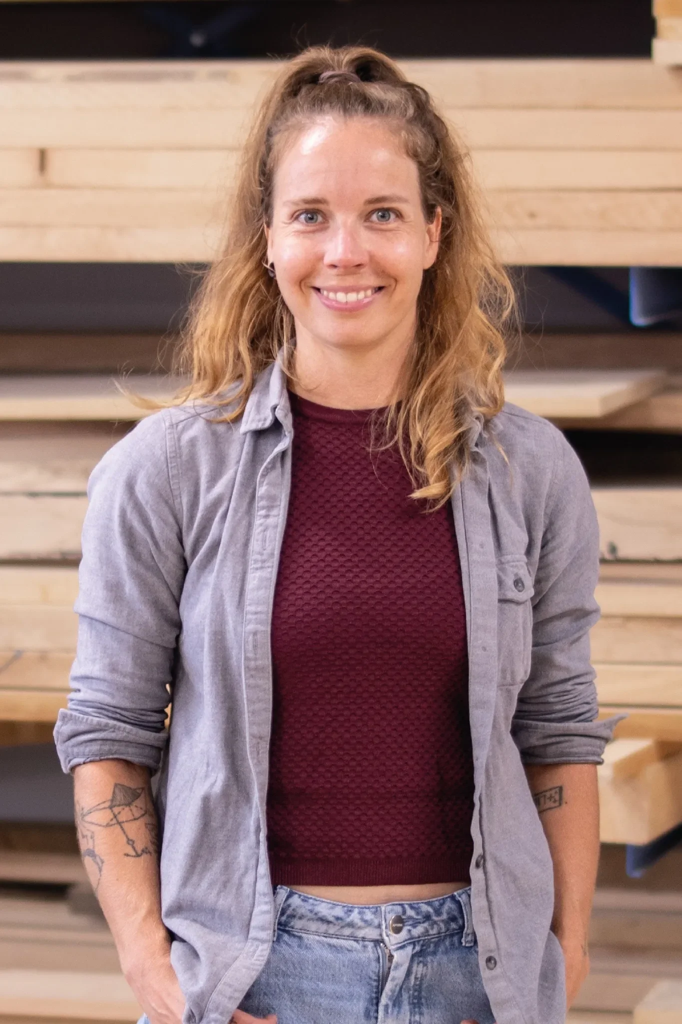 Eine Frau mit lockigem, blondem Haar, die vor einem Regal mit Holz in einem Holzlager steht, trägt eine graues Flanell-Hemd, ein dunkelrotes Oberteil und Jeans, und blickt lächelnd in die Kamera.