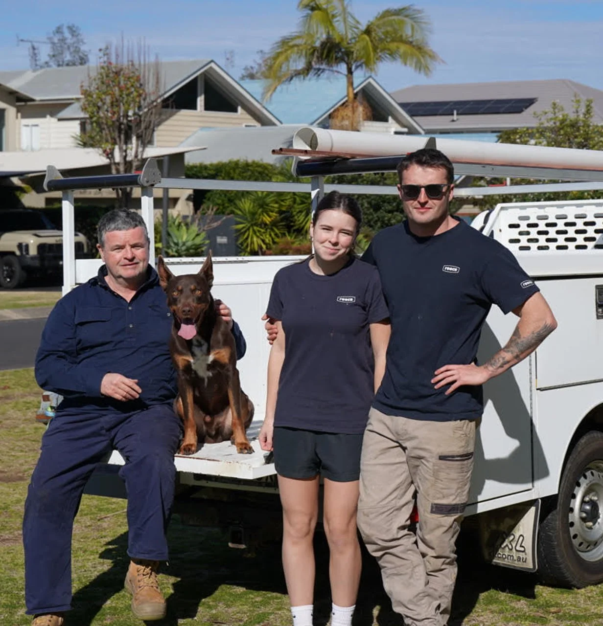 Three people and a dog pose in front of a utility vehicle with equipment, on a residential street with houses, trees, and a palm tree in the background.