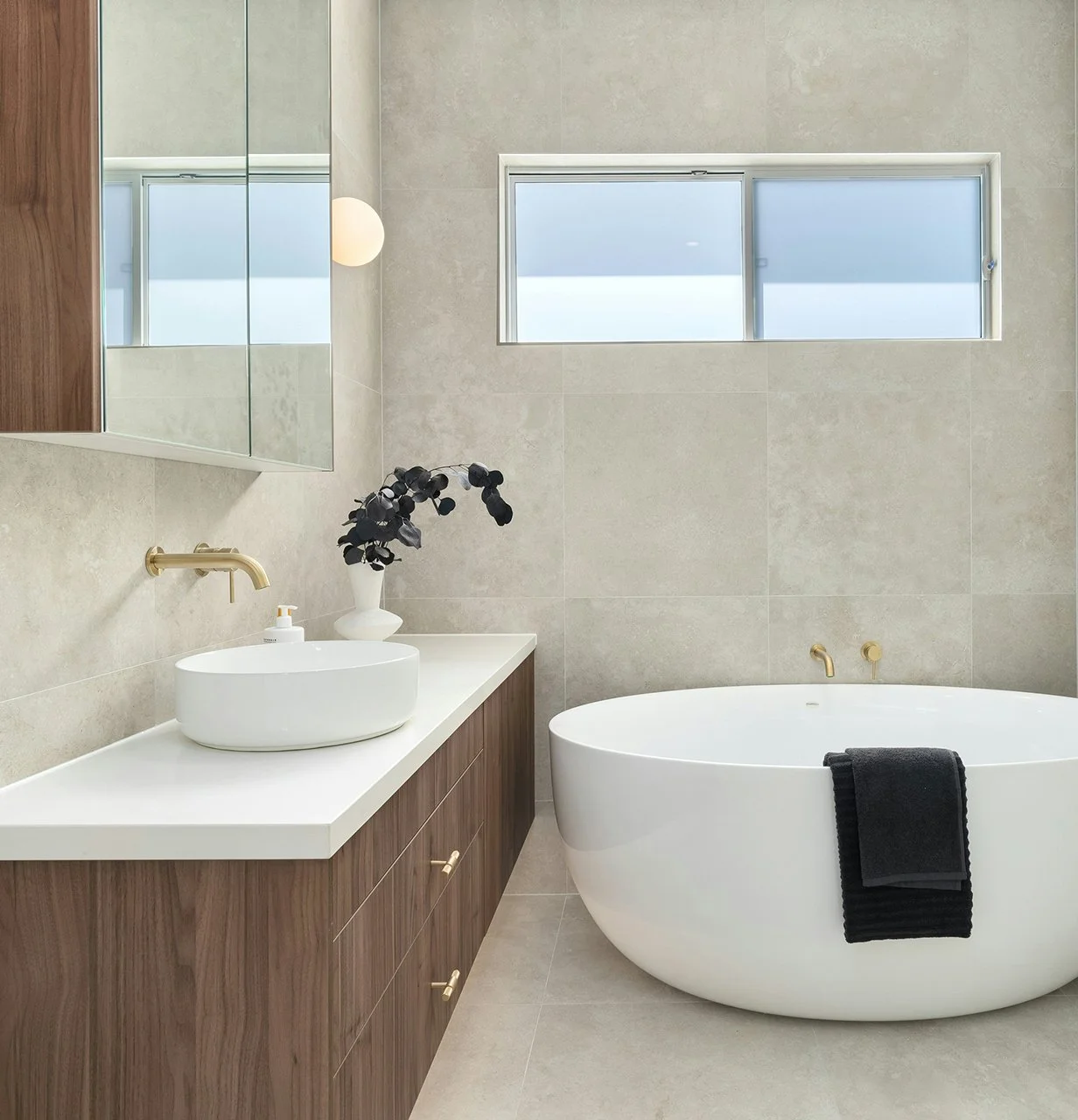 Modern bathroom with beige tile walls, a white bathtub with a black towel, wooden vanity with a white vessel sink, gold fixtures, and a window with frosted glass.