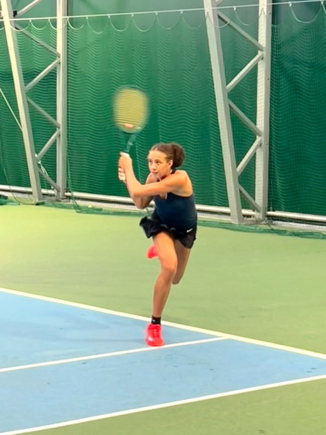 Bucks County Player playing tennis on an indoor court, preparing to hit a tennis ball with a racket, wearing a black athletic outfit and bright red athletic shoes.