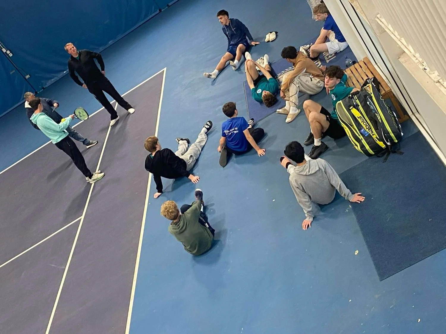 Bucks County Tennis Players sitting and lying on the floor of an indoor tennis court, with a coach or instructor standing nearby holding a tennis racquet and giving instructions.