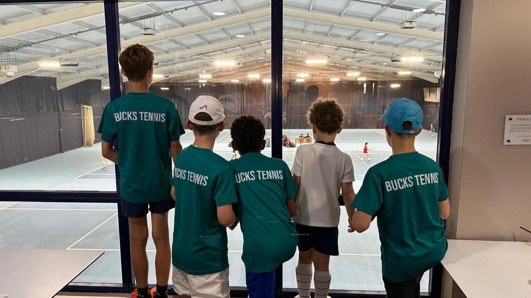 Bucks team of five boys with Bucks County tennis uniforms, some wearing caps, watching a tennis match through a glass window at an indoor tennis facility.