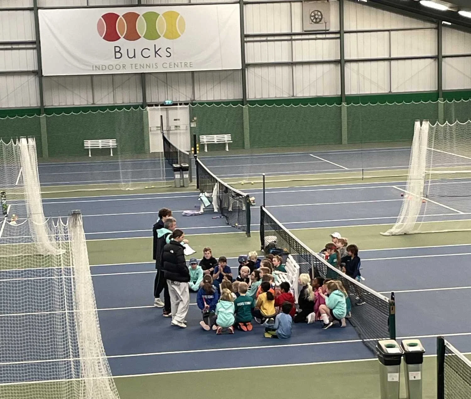 Bucks County Tennis players sitting on the floor of an indoor tennis court, listening to a coach or instructor standing nearby.