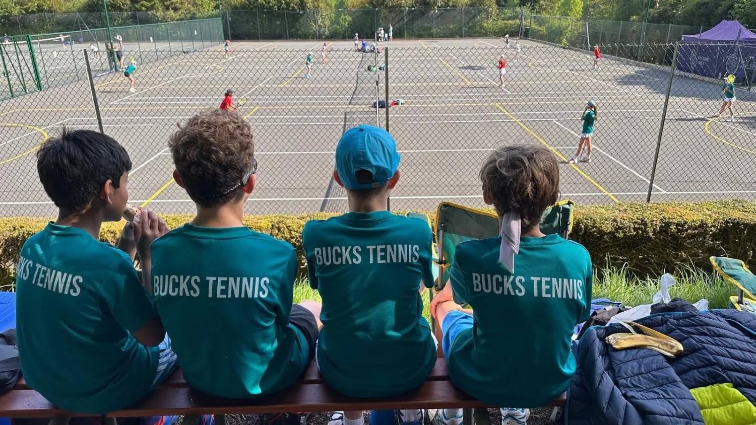 Four children wearing teal shirts with 'BUCKS TENNIS' printed on the back, sitting on a bench and watching a tennis practice on outdoor courts.
