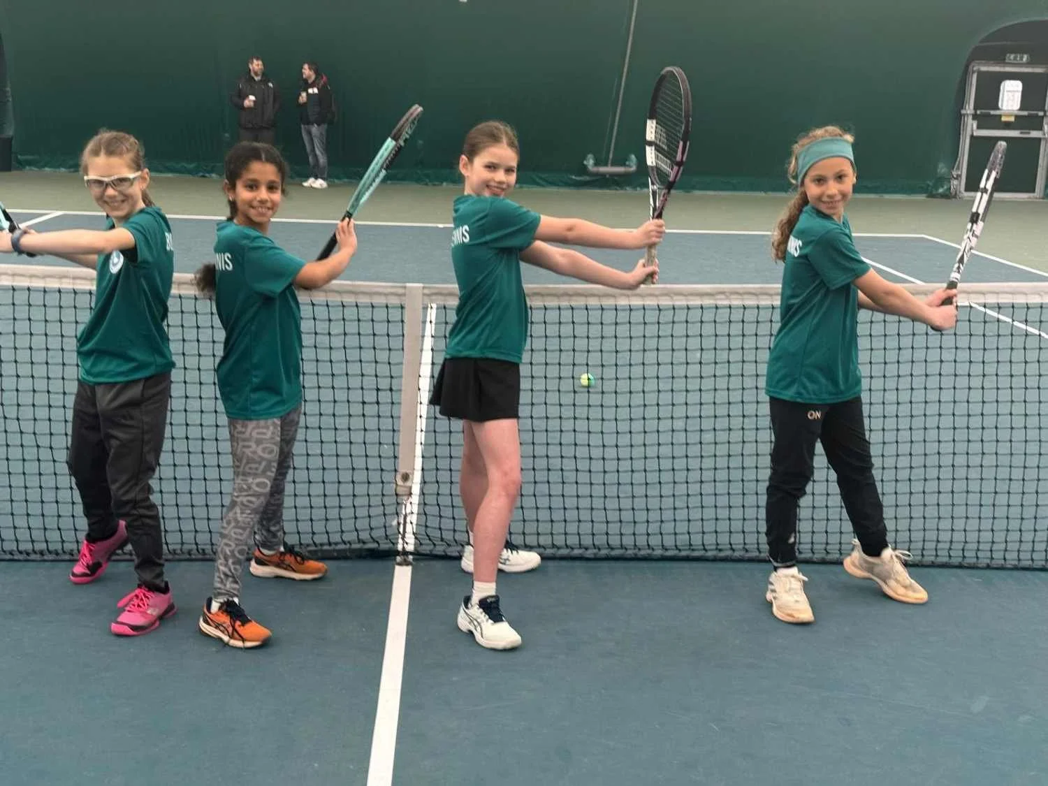 Under 8 Bucks County Tennis Players standing in a line on a tennis court, holding tennis rackets and smiling, wearing matching green uniforms.