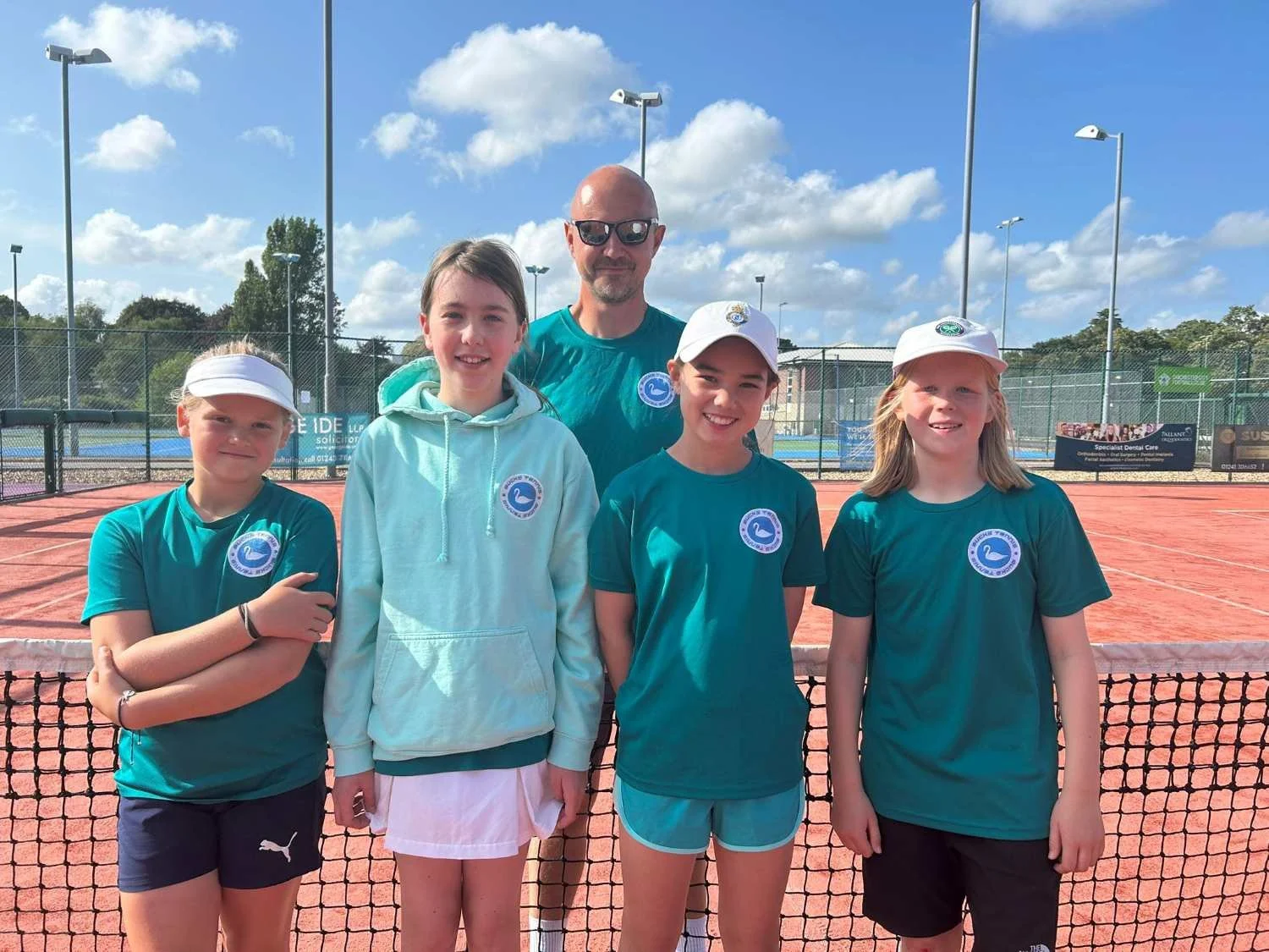 Bucks County Tennis players and their male coach standing on a tennis court, smiling, wearing teal shirts with a circular logo, two girls with white visors, and a sunny day with a bright blue sky and scattered clouds.