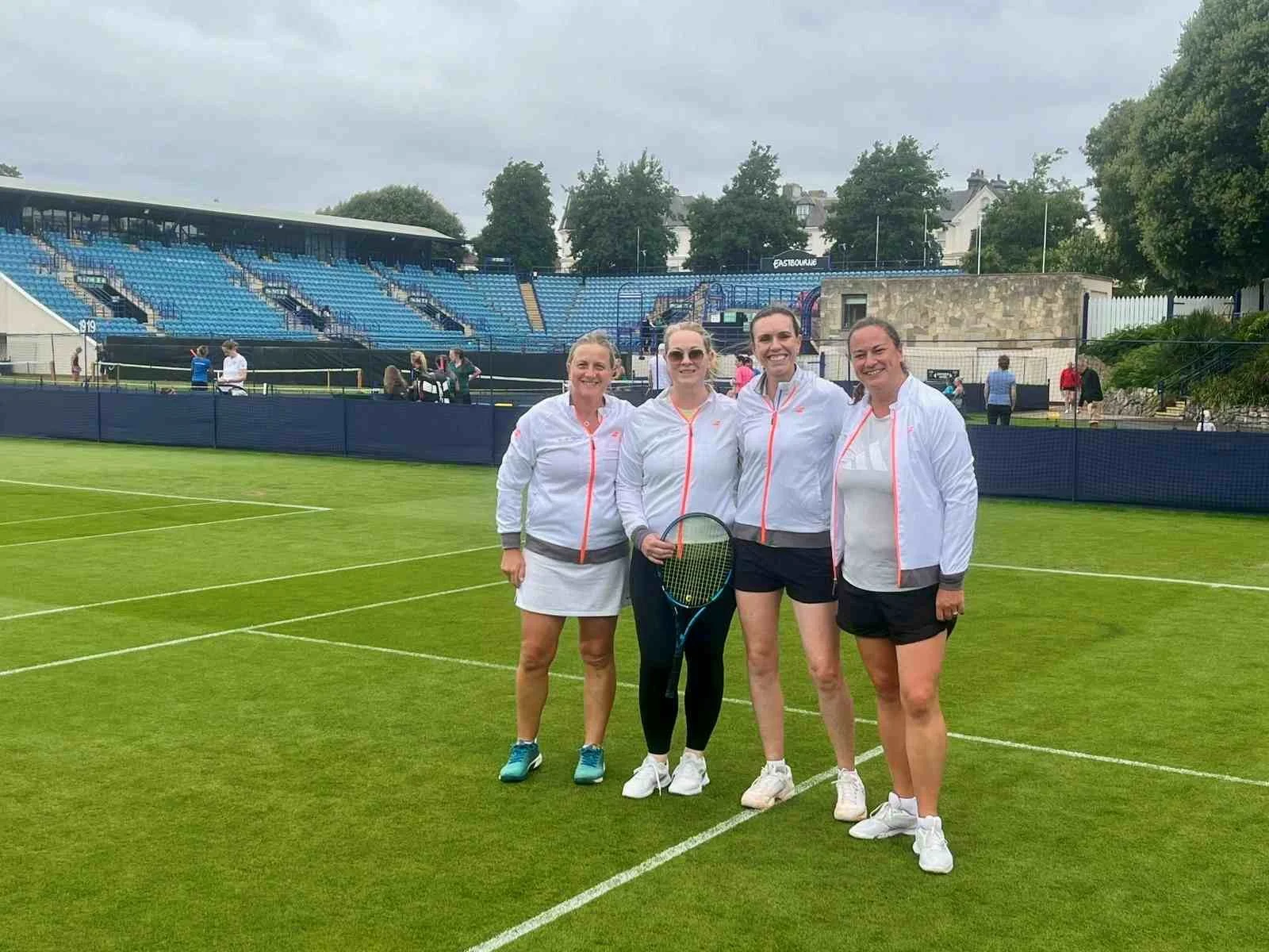 Bucks over 35 county tennis team standing on a tennis court in Eastbourne, dressed in sporty white jackets and shorts, holding tennis rackets, smiling at the camera with stadium seats and trees in the background.