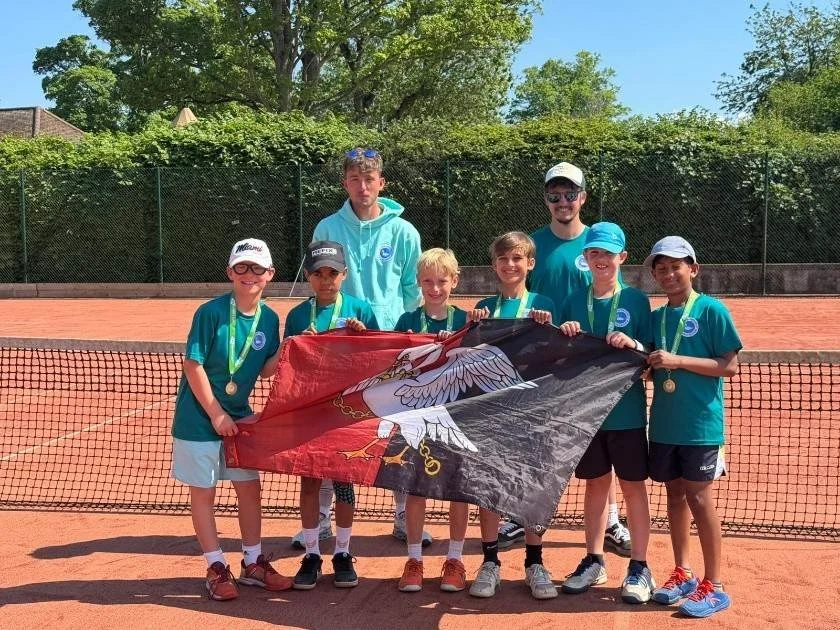 Bucks County tennis players and coaches on a clay tennis court holding a flag, celebrating a victory, with trees and a fence in the background.