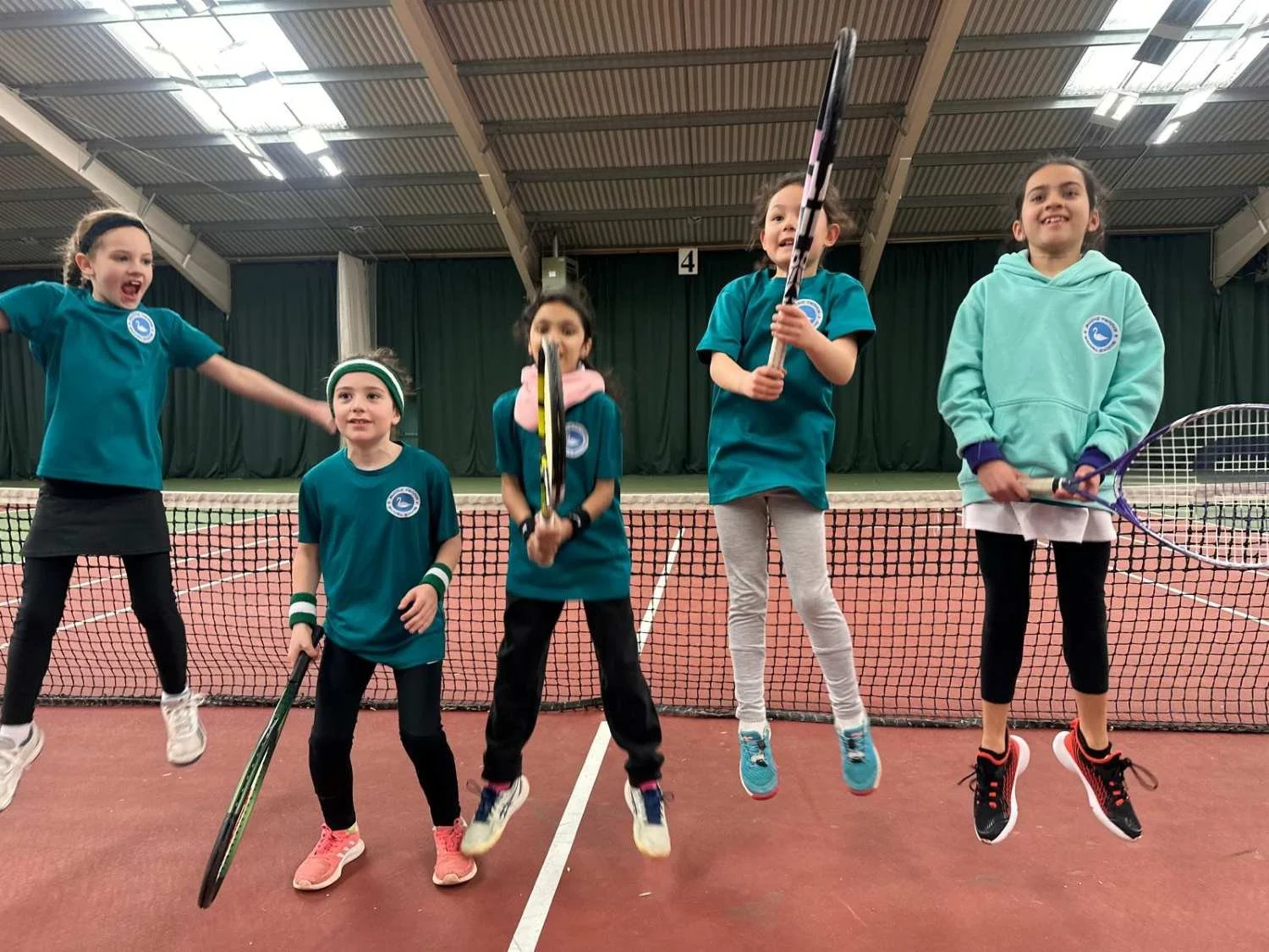 Under 8 bucks county tennis players on an indoor tennis court, wearing teal sports shirts with a tennis logo, jumping mid-air and holding tennis rackets, smiling and having fun.
