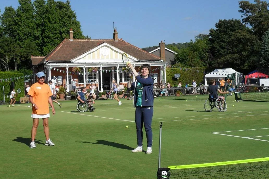 People playing tennis on an outdoor court with a house in the background on a sunny day.