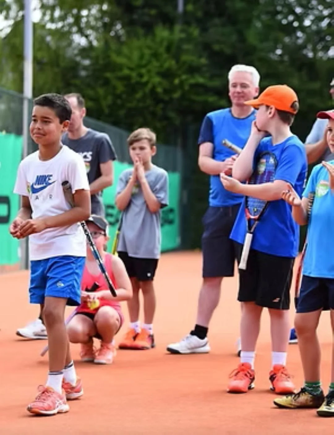 Children and a coach on a tennis court, with some kids holding tennis rackets, preparing for a game.