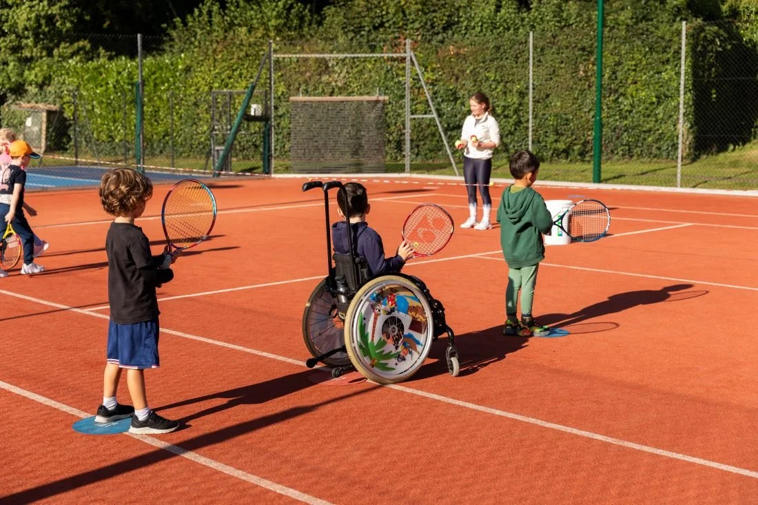 Children playing tennis on a court with a coach, including a child in a wheelchair, during daytime.