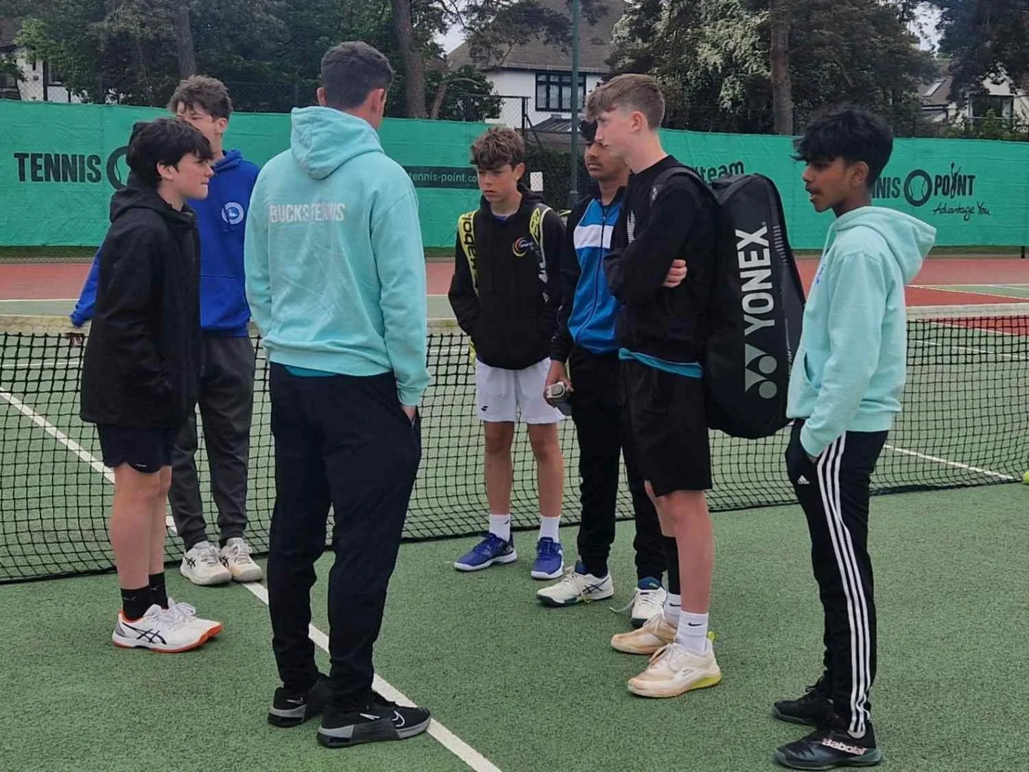 Bucks County Tennis players standing on a tennis court, engaged in a discussion, with tennis equipment and a green background.