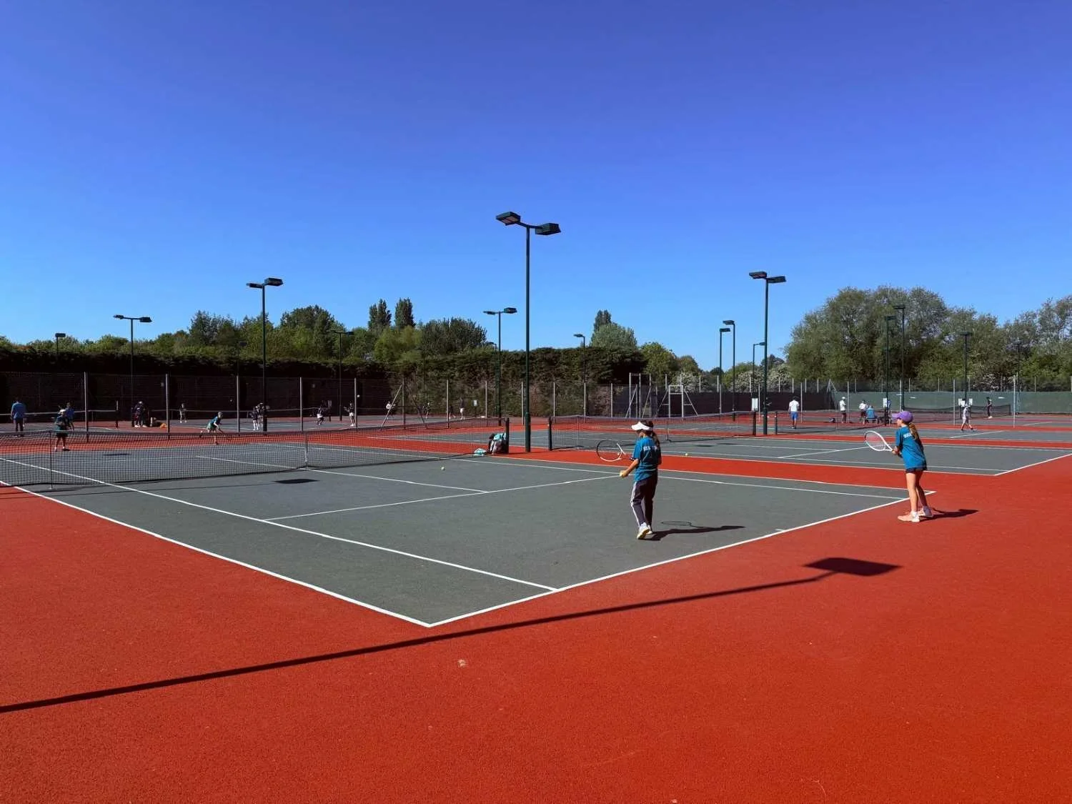 Bucks County players playing tennis on outdoor tennis courts with trees in the background and a clear blue sky.