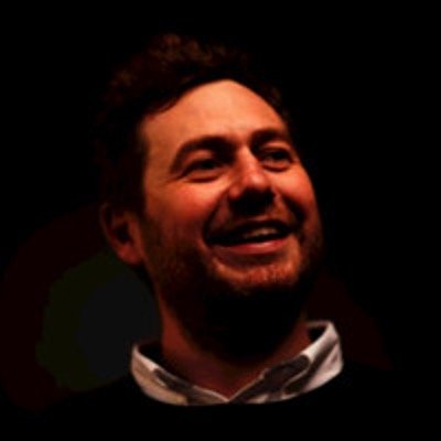 Smiling woman with shoulder-length black hair wearing a blue top, standing among green plants.