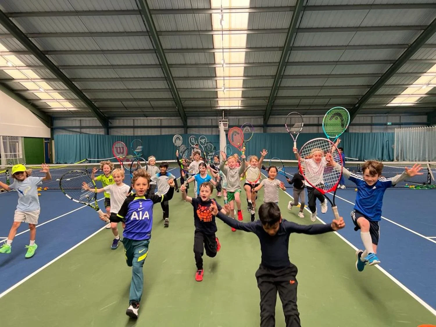 Bucks County Players playing tennis indoors on a court with a high ceiling, holding tennis rackets, some jumping and smiling.