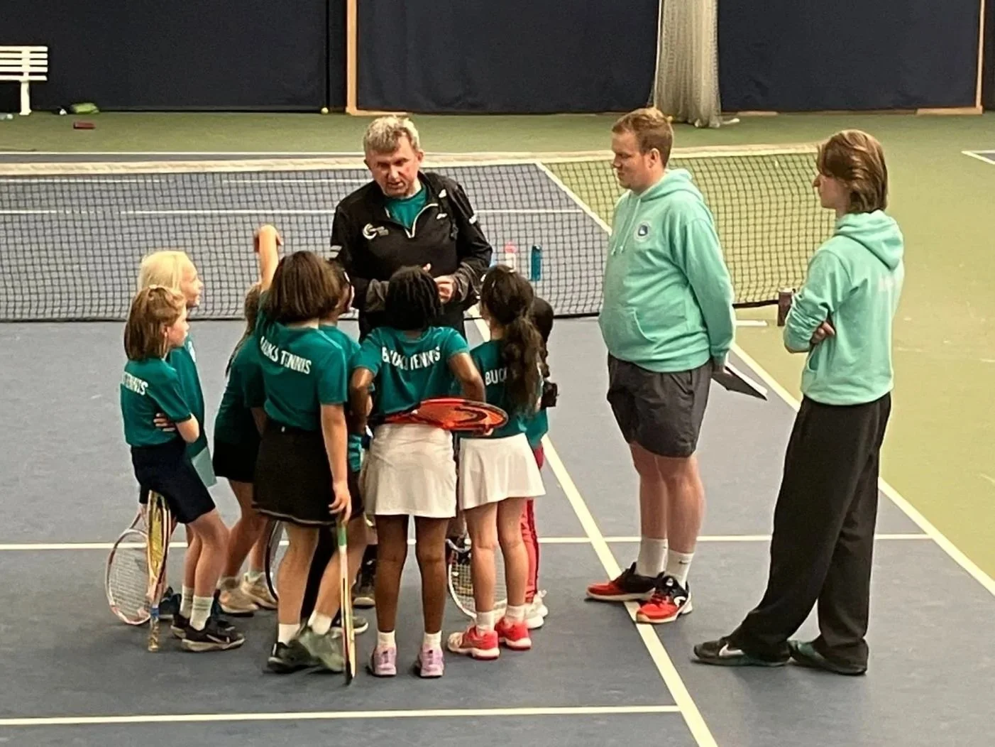 Under 8 junior bucks county players and their coaches, Neale Proud, on an indoor tennis court, engaged in a discussion during a tennis practice or lesson.