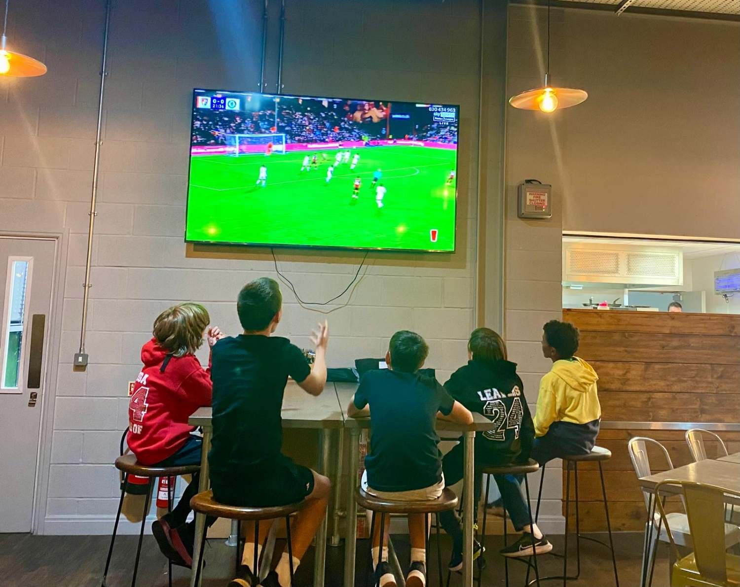 Bucks County Tennis players sitting on high stools at a bar-style table watching a soccer game on a large wall-mounted TV, in a dimly lit cafe or bar with modern decor.