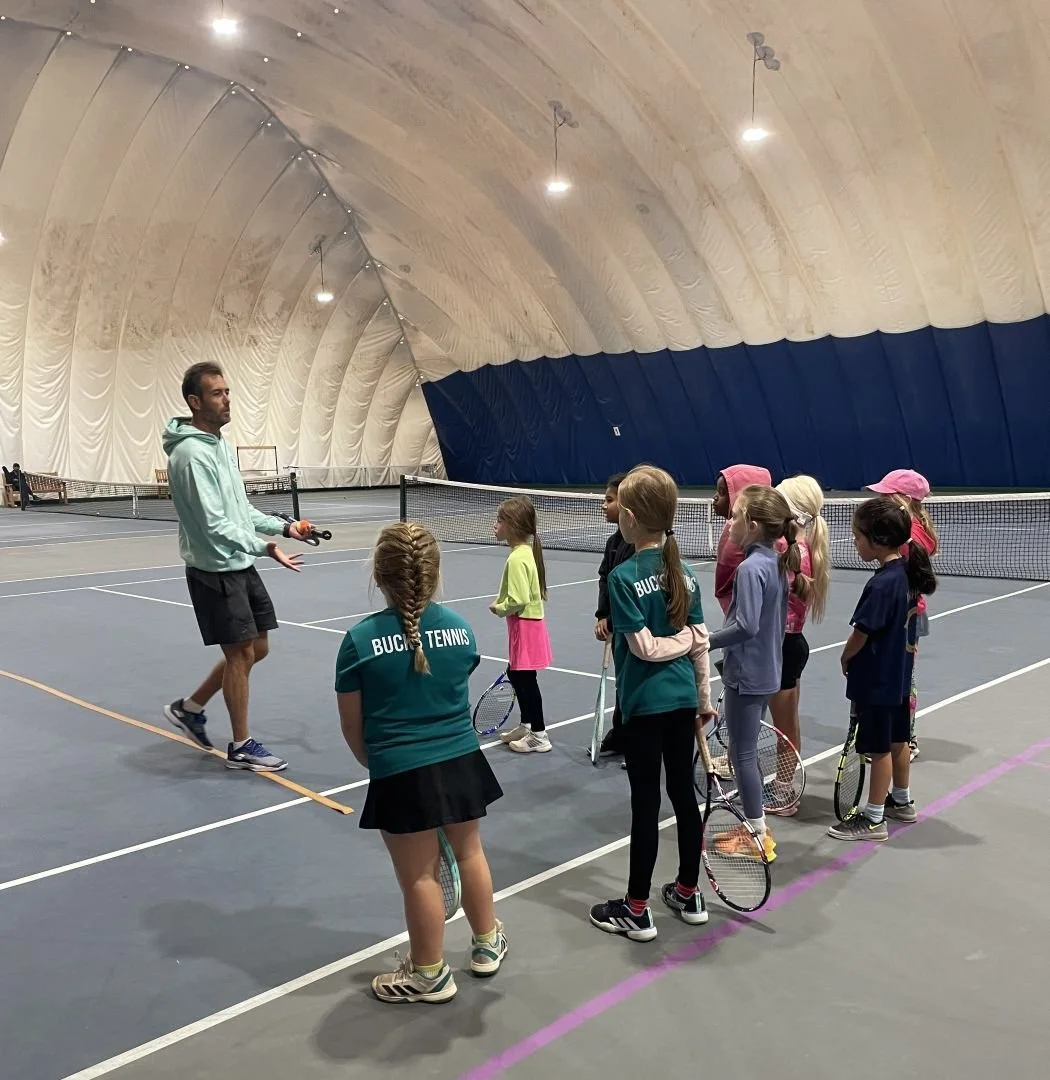 A bucks county tennis coach teaching a group of young girls inside an indoor tennis court.