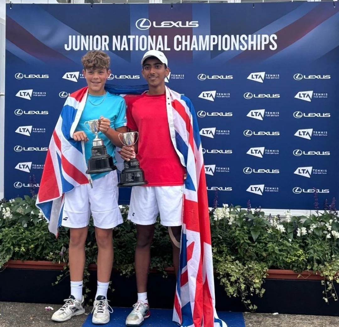 Two Buckinshamshire tennis players celebrating at a tennis championship, holding two trophies, wearing Union Jack flags around their shoulders, standing in front of a blue backdrop with logos and the text 'Lexus Junior National Championships.'