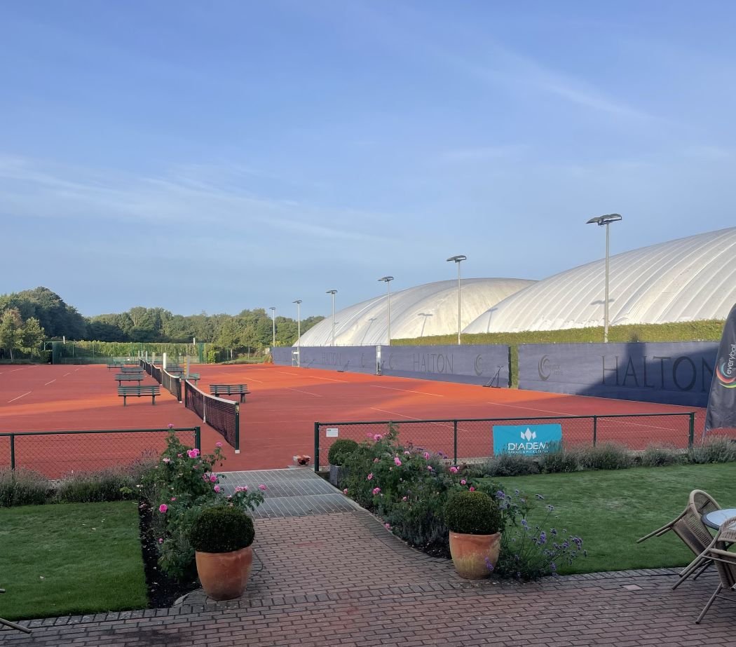 Halton Tennis Centre with a red clay surface, surrounded by a black fence, with benches and trees in the background. There is a pathway with potted flowers leading to the court and a blue advertisement banner.