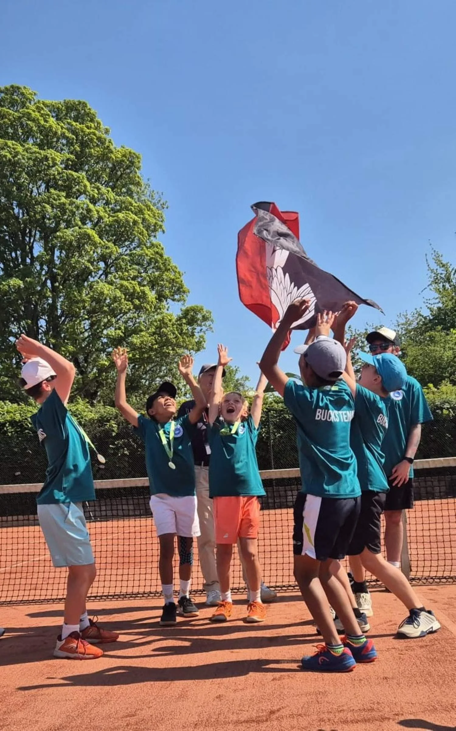 Bucks County Tennis players celebrating on a tennis court, raising hands and holding a flag, with trees and a clear blue sky in the background.
