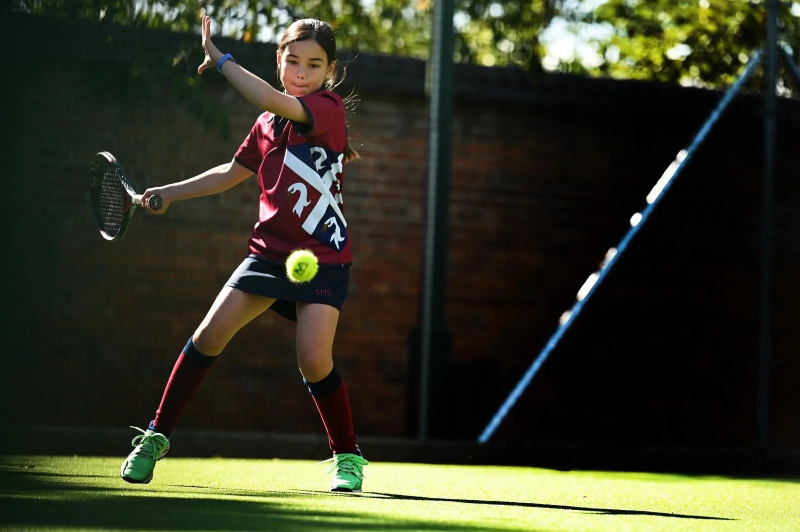 A man with a dark skin tone, wearing a gray shirt, gray shorts, and a blue cap, watching a tennis practice with a basket of tennis balls nearby on an outdoor tennis court. A woman with a light skin tone, wearing a white tank top, black shorts, and a bright green cap, holds a tennis racket and stands behind the net.