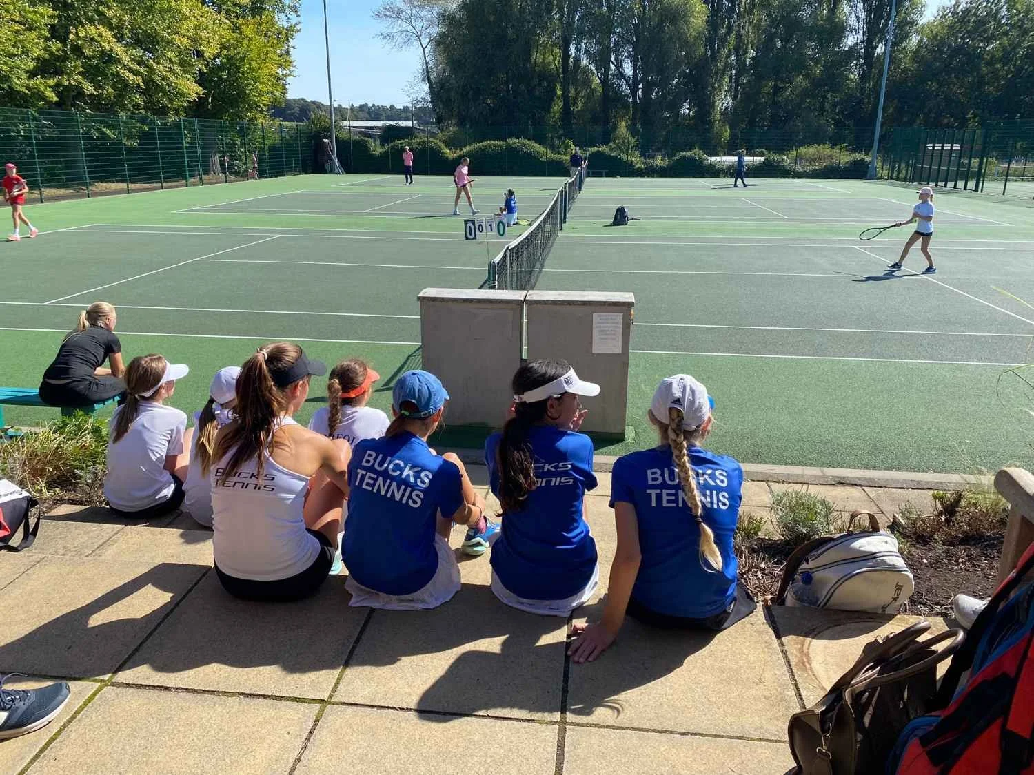 Bucks County Tennis Players watching a tennis match from the sidelines of an outdoor tennis court.
