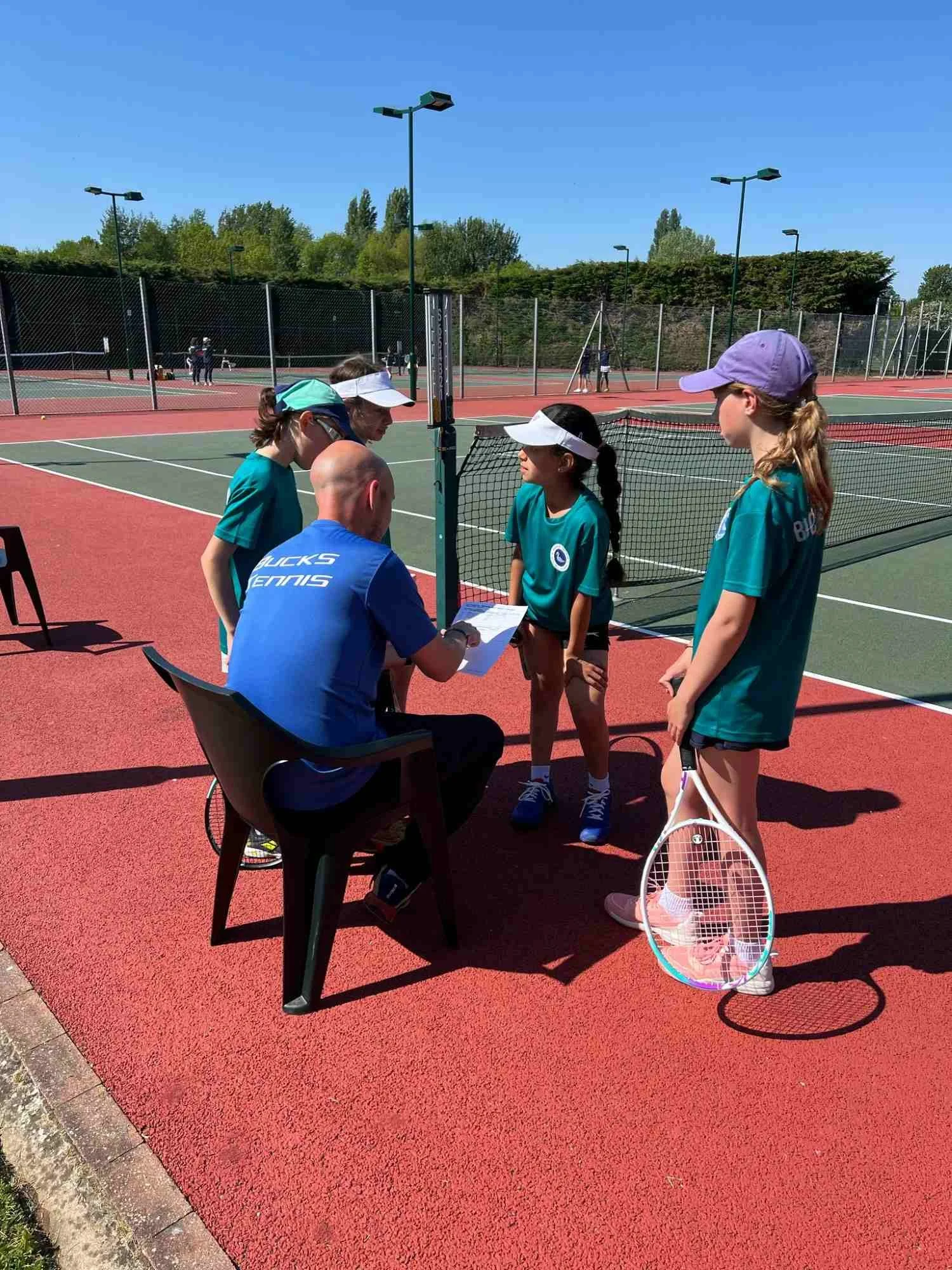 Bucks County Girls in tennis uniforms receiving instructions from a coach on a tennis court during daytime. The court is outdoors with multiple courts and green trees in the background.