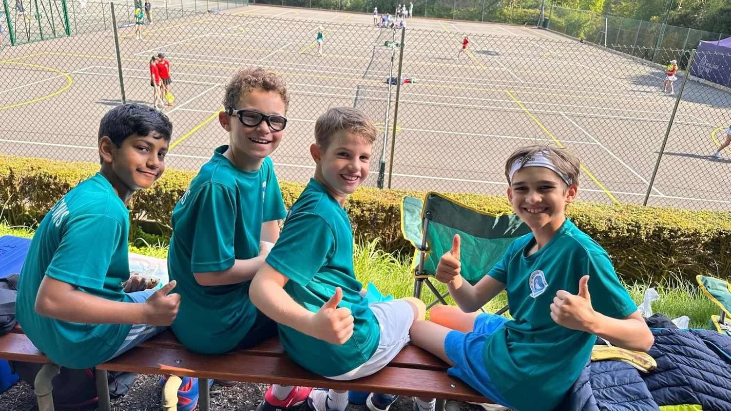 Bucks County Tennis boys sitting on a bench at an outdoor tennis court, smiling and giving thumbs up, with other children playing tennis in the background.