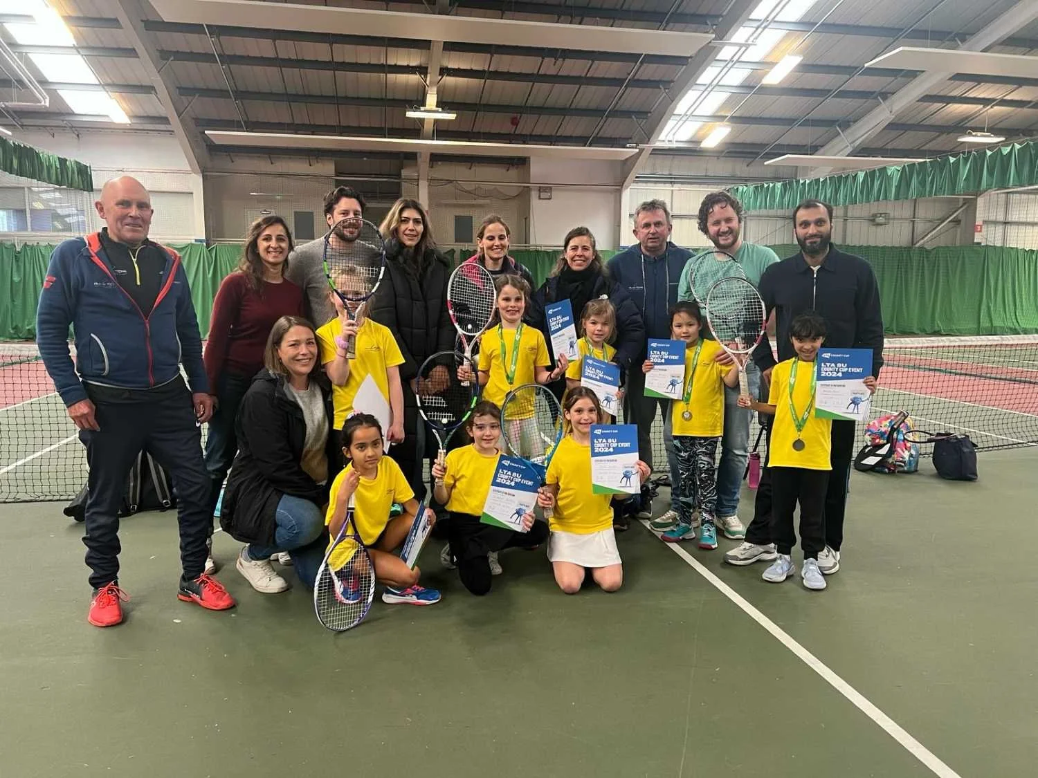 Group photo of Bucks County Tennis Players and parents on an indoor tennis court, with children holding tennis rackets and certificates, some wearing medals, celebrating a tennis event.